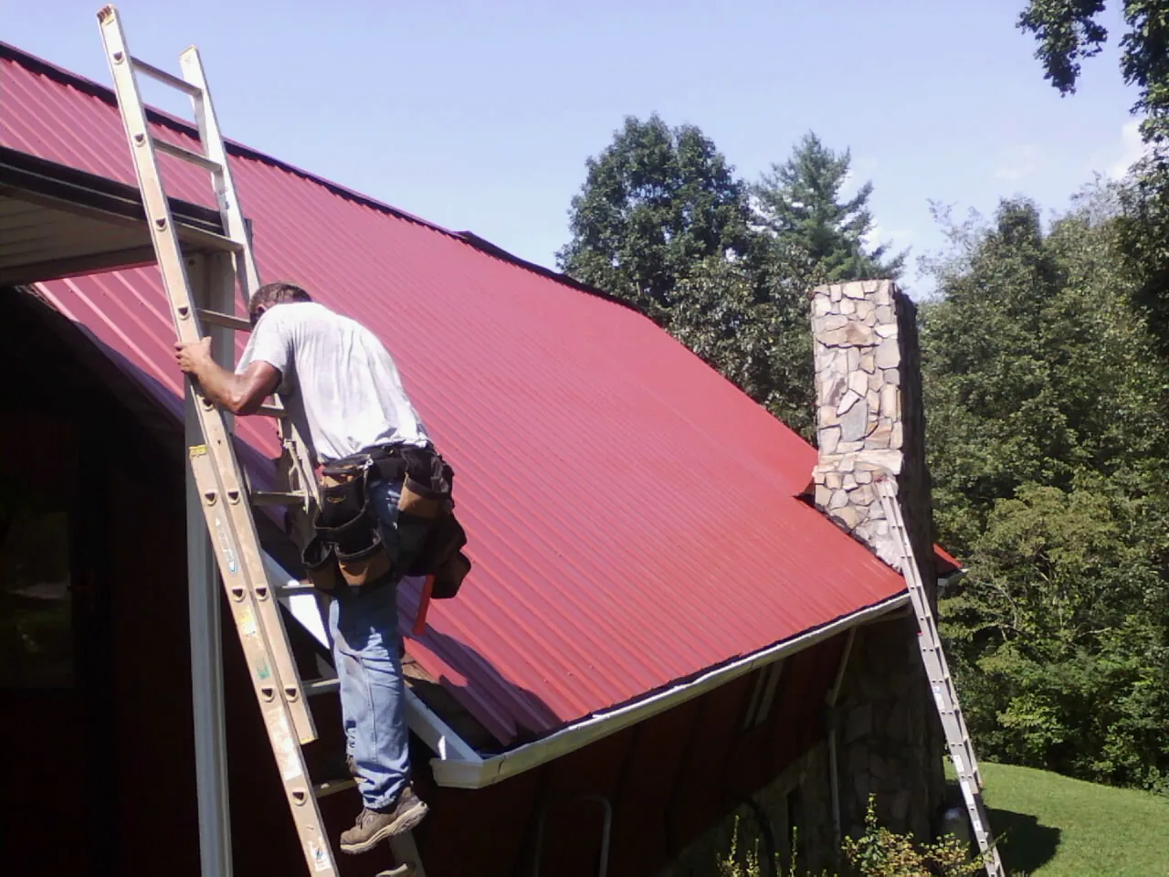 Roofer on a ladder beside a red metal roof and stone chimney — Soffit & Fascia Repair by Roofing Brothers in Sharps Chapel